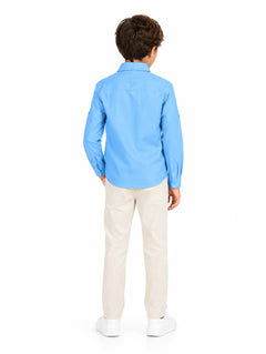 Niño modelando una camisa azul de manga larga, pantalones beige y tenis blancos sobre fondo blanco.